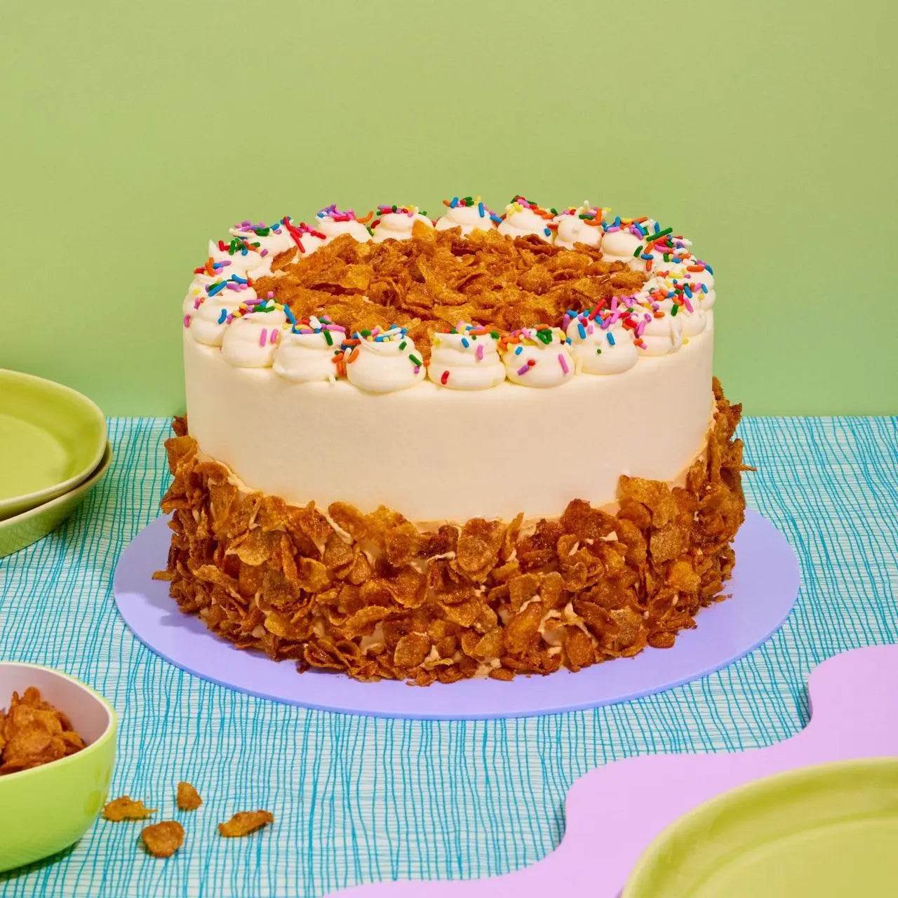 Cereal Milk Cake by Crumbs & Doilies with cereal clusters, sprinkles, and smooth buttercream on a purple board.