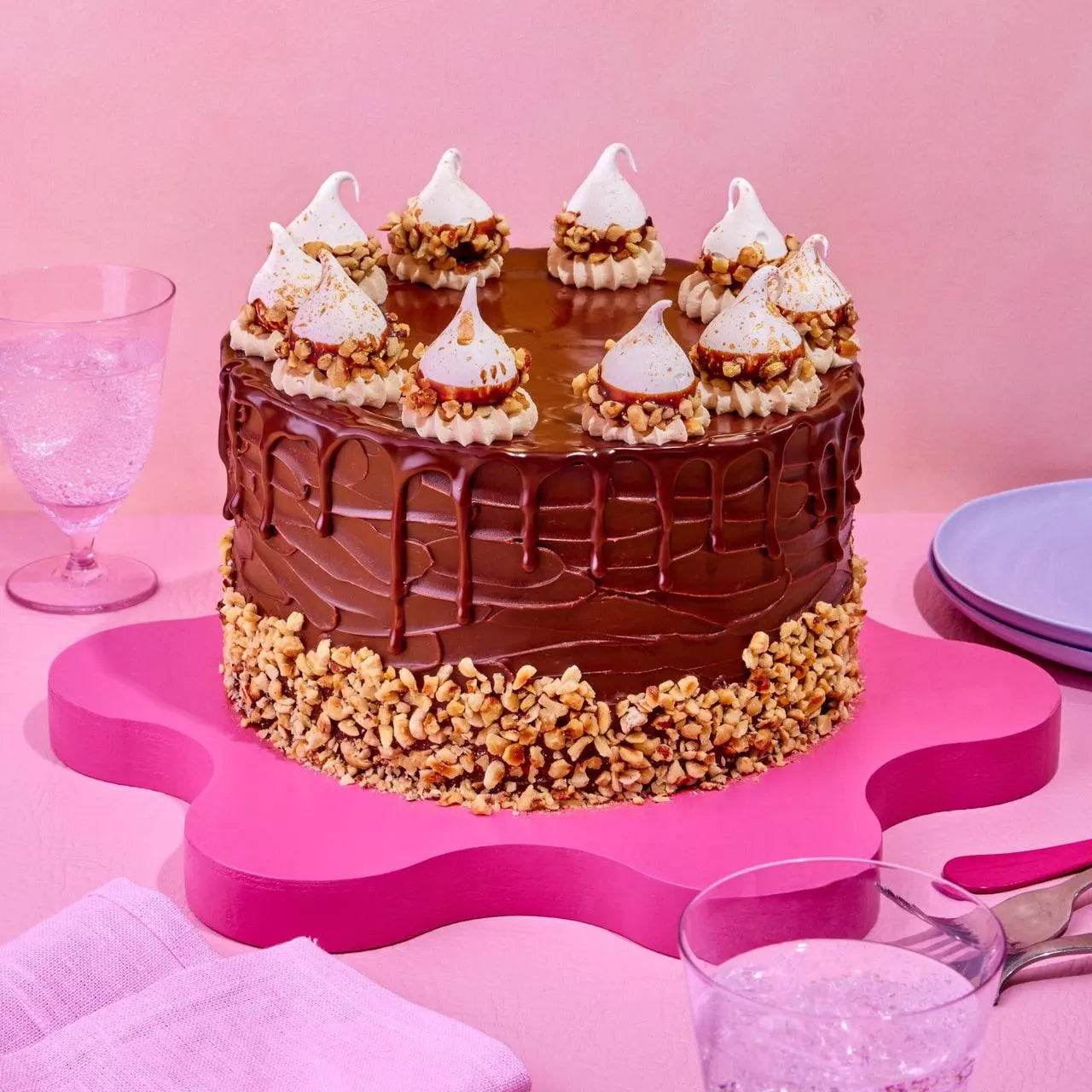 Ferrero Rocher Cake with chocolate ganache drip, gold-flecked meringues, and chopped hazelnuts on pink board by Crumbs & Doilies