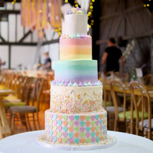 Multicolored tiered cake on a table with a blurred background of chairs and people.