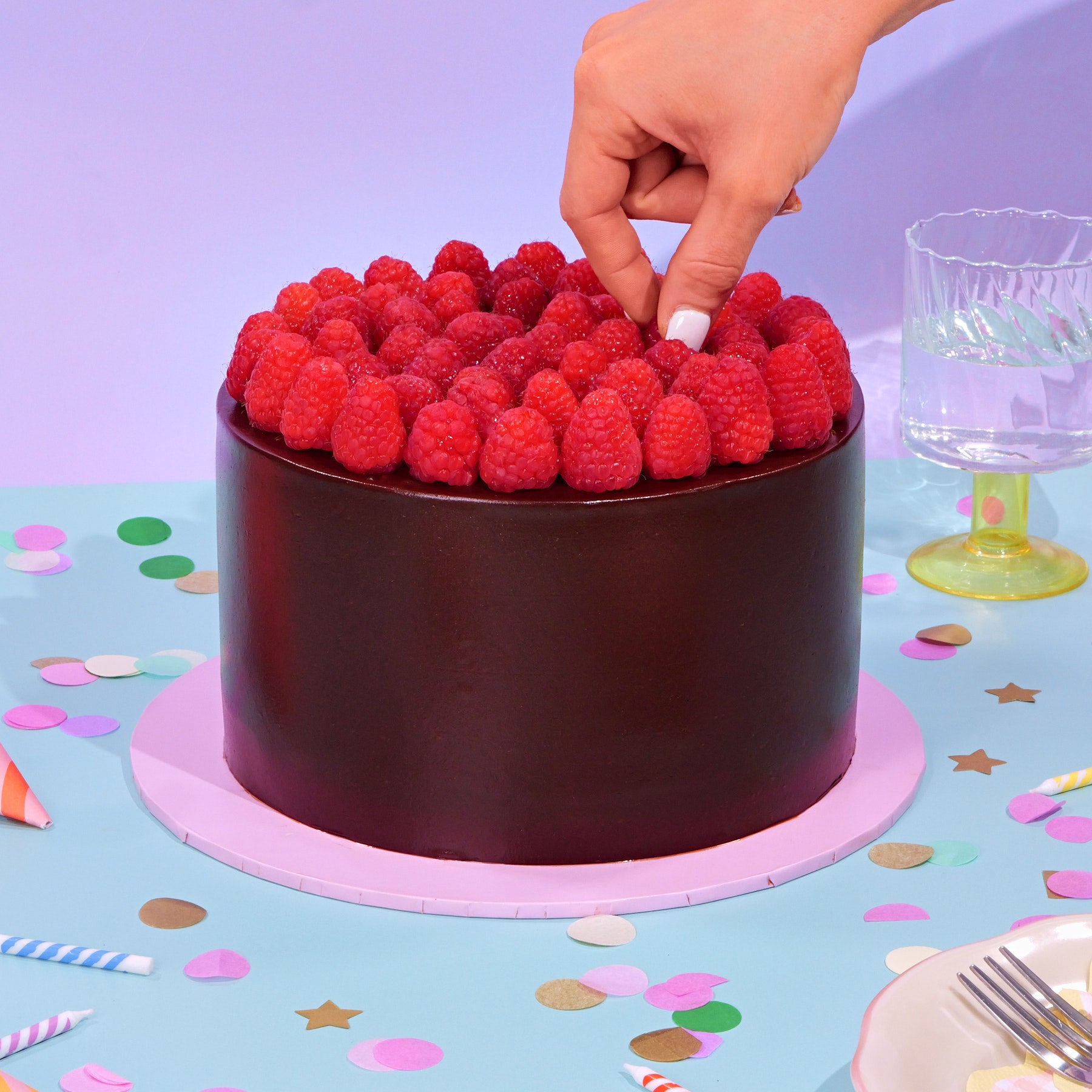 Chocolate & Raspberry Cake topped with fresh raspberries on smooth dark chocolate frosting with party decor nearby.
