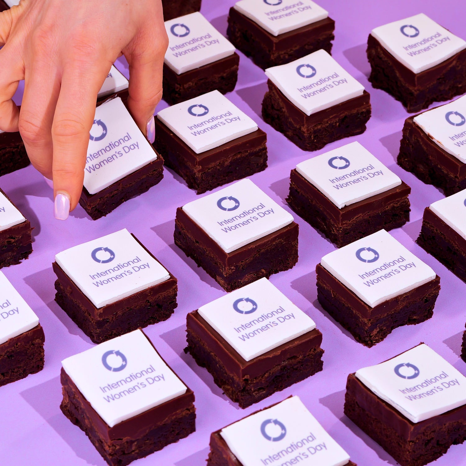 International Women's Day Brownies with Edible Logos featuring ganache topping arranged on a purple surface with hand lifting one brownie