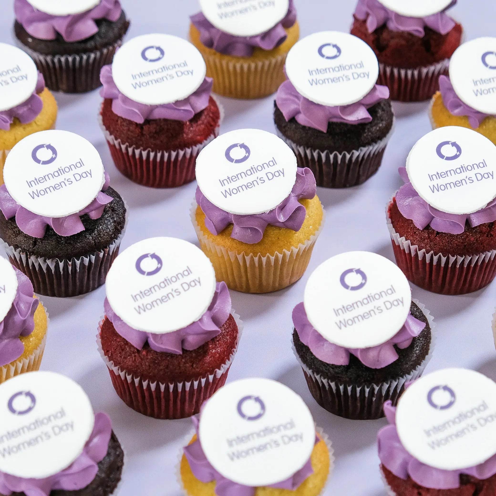 International Women's Day Cupcakes by Crumbs & Doilies with purple icing and edible logo toppers.