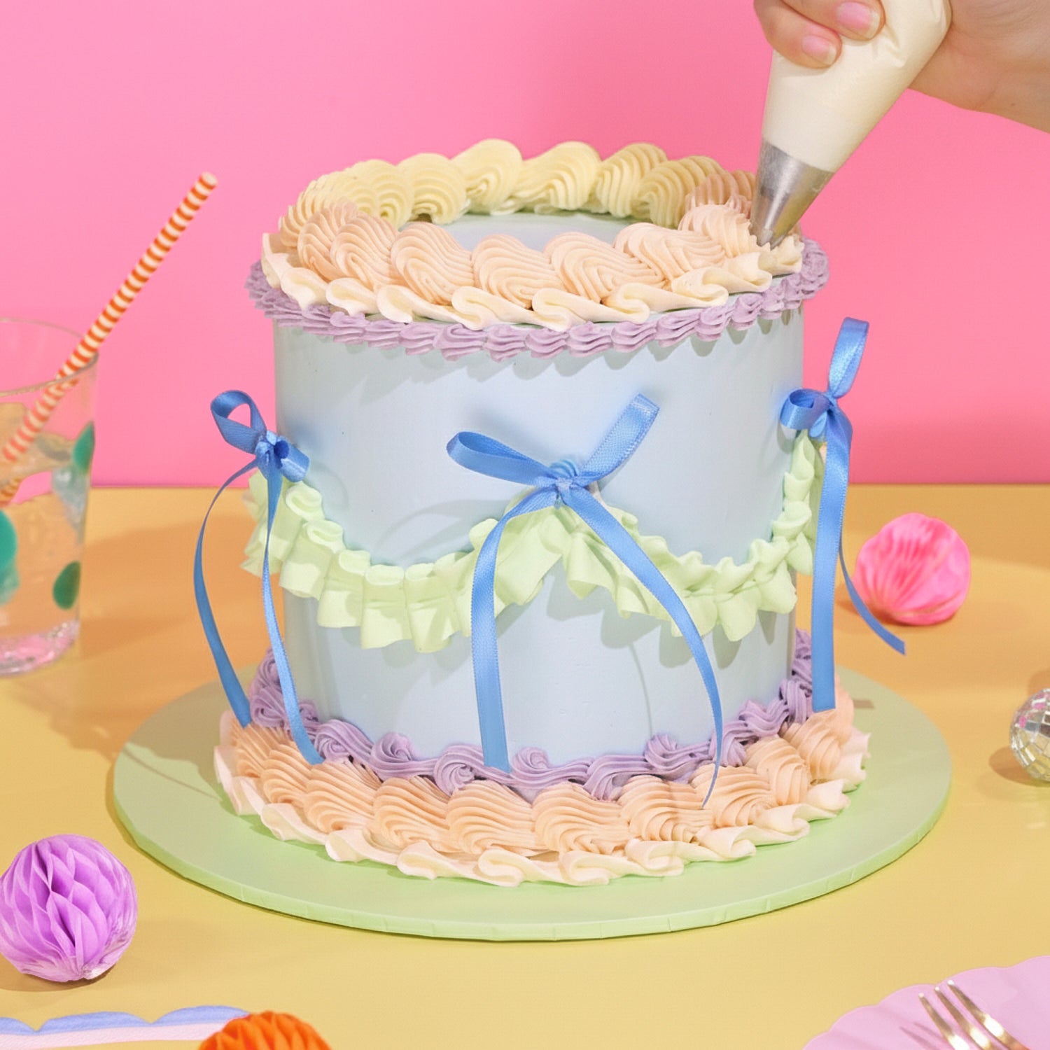 Hand decorating a pastel Bespoke Vintage Cake with piped icing ruffles and blue ribbon bows on a yellow surface.